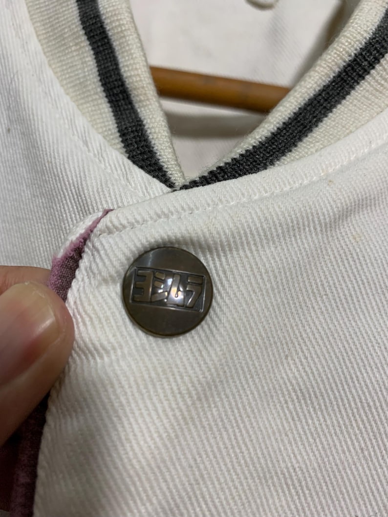 May include: Close-up of a white jacket with a button. The button is bronze-colored with Japanese text. The jacket has a striped collar in white, gray, and black. The edge of the jacket is lined with a pink fabric.