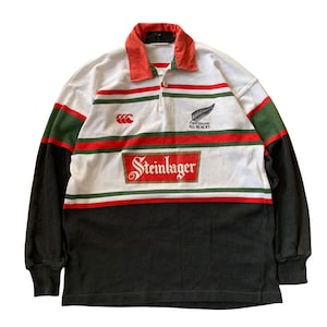 May include: A long-sleeve rugby shirt featuring a white base with horizontal red and green stripes. The shirt has a red collar, black sleeves, and a black lower section. It displays the Steinlager logo, the Canterbury logo, and the All Blacks fern.