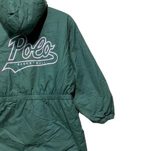 Pu&ograve; includere: Giacca con cappuccio verde scuro con la scritta "Polo" in corsivo bianco e la scritta "Basketball" sotto. La giacca ha una vita con coulisse e una cerniera a tutta lunghezza. La giacca &egrave; realizzata in un materiale leggero.