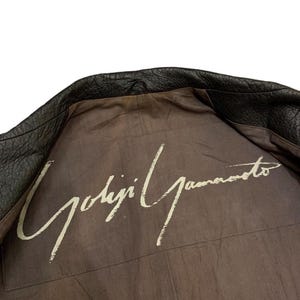May include: Black leather jacket with the interior lining visible. The lining is a muted brown color with the white signature "Yohji Yamamoto" printed across the back.