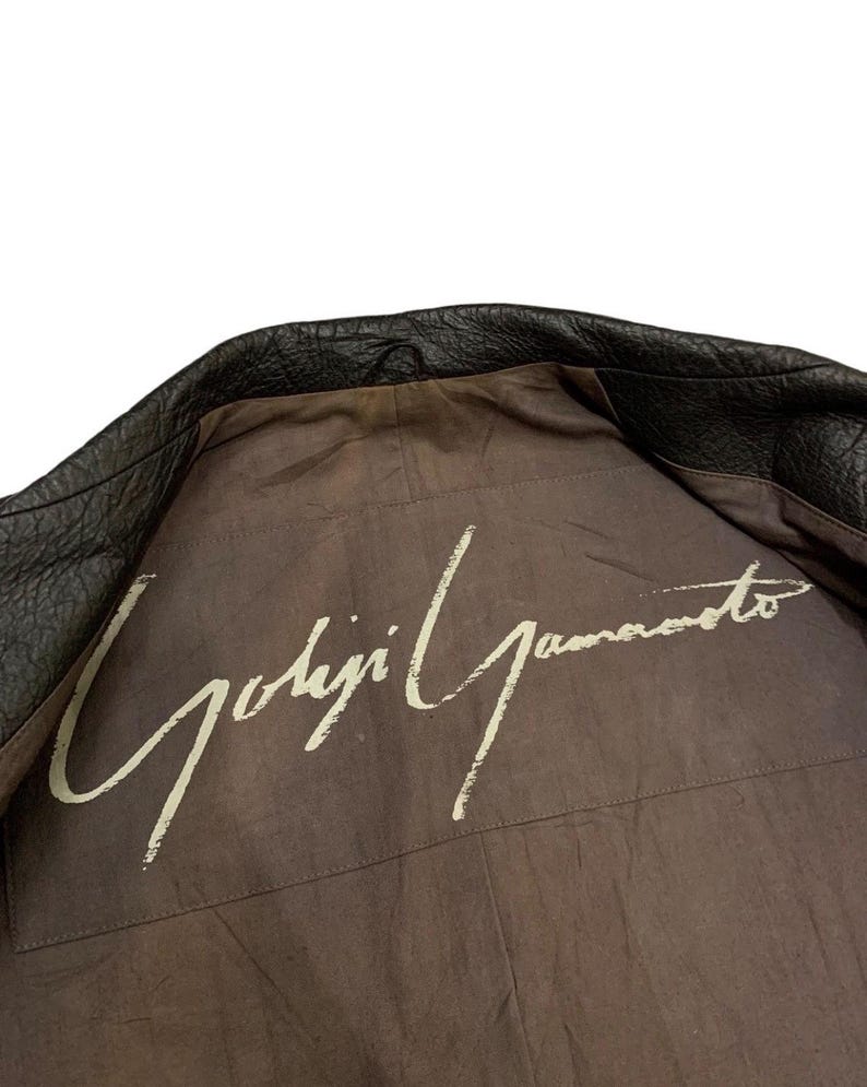 May include: Black leather jacket with the interior lining visible. The lining is a muted brown color with the white signature "Yohji Yamamoto" printed across the back.