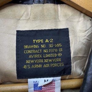 May include: Close-up of a black label from an A-2 jacket, with text including "TYPE A-2", "AVIREX LIMITED", and "U.S. ARMY AIR FORCES". A smaller tag shows the American flag and "AVIREX LTD". The jacket's interior is visible.