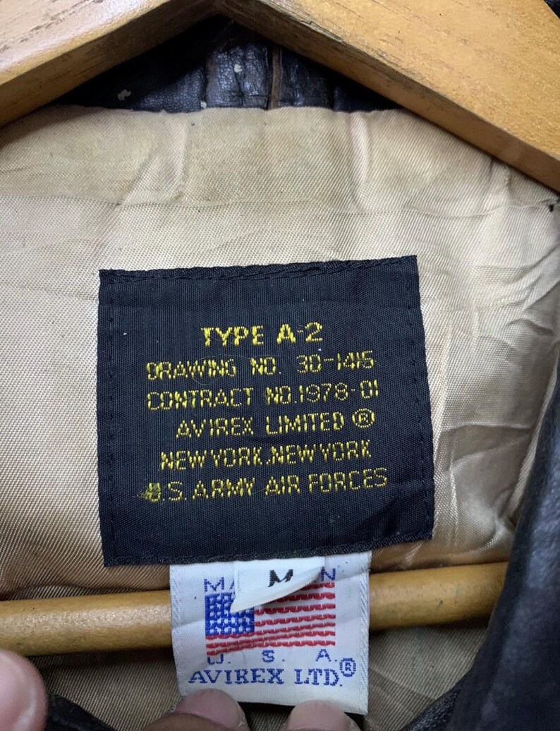 May include: Close-up of a black label from an A-2 jacket, with text including "TYPE A-2", "AVIREX LIMITED", and "U.S. ARMY AIR FORCES". A smaller tag shows the American flag and "AVIREX LTD". The jacket's interior is visible.