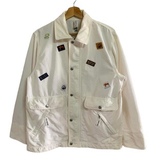 Puede incluir: Una chaqueta blanca con varios parches, incluyendo un zorro, un oso y una escena de montaña. La chaqueta tiene cierre de botones y dos bolsillos grandes.