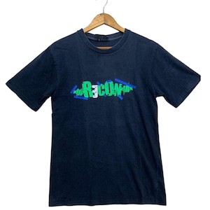 May include: Dark navy blue t-shirt with the word "R3COW" in green, blue, and white graffiti-style lettering across the chest. The short-sleeved shirt is hanging on a wooden hanger against a white background.