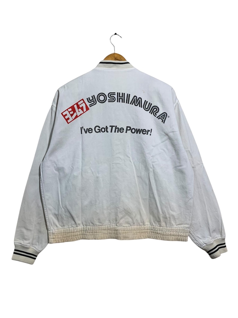 May include: White bomber jacket with the text "YOSHIMURA" in black and red, and the phrase "I've Got The Power!" The jacket has black and white striped cuffs and collar.