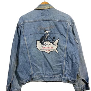 May include: Light blue denim jacket featuring the Wrangler logo and a graphic of a cowboy riding a horse on the back. The graphic is set against a map of the USA. The text "American Top Cowboys Choose The Wrangler" is also visible.