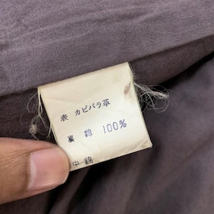 May include: A close-up of a clothing tag with Japanese text and the number 100%. The tag is rectangular, with a white background and black text. The fabric surrounding the tag is a muted purple color.