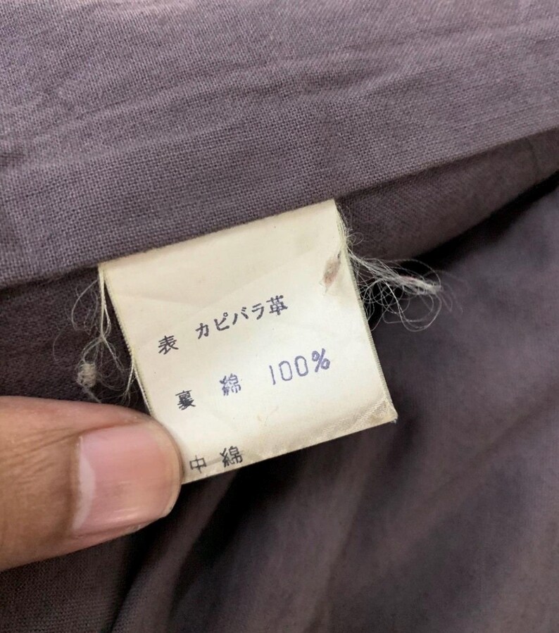 May include: A close-up of a clothing tag with Japanese text and the number 100%. The tag is rectangular, with a white background and black text. The fabric surrounding the tag is a muted purple color.