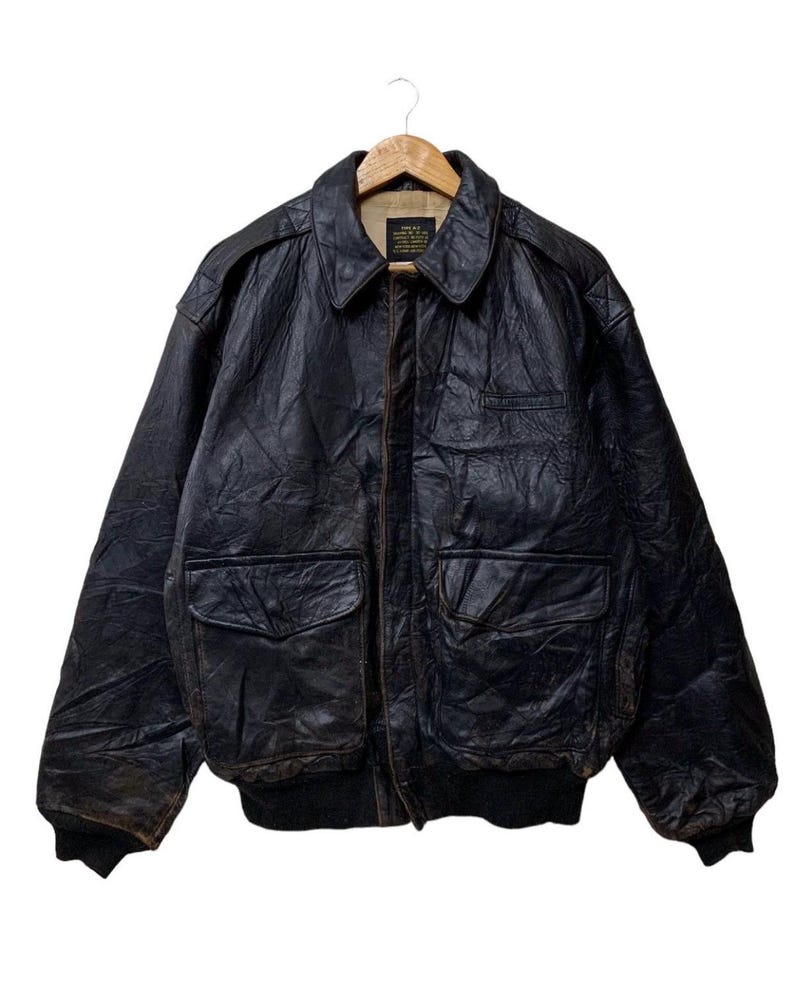 May include: A vintage black leather bomber jacket with a collared neckline, two front pockets, and a ribbed hem and cuffs. The jacket is hanging on a wooden hanger against a white background. The leather has a distressed appearance.