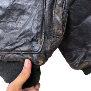 May include: Close-up of a vintage black leather bomber jacket. The jacket shows signs of wear with a distressed appearance, including creasing and discoloration. The ribbed cuffs and collar are black.