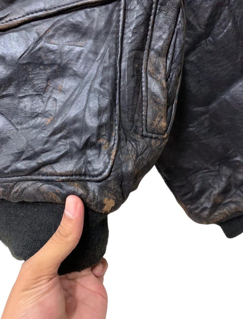 May include: Close-up of a vintage black leather bomber jacket. The jacket shows signs of wear with a distressed appearance, including creasing and discoloration. The ribbed cuffs and collar are black.