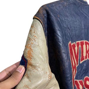 Puede incluir: Chaqueta universitaria vintage azul y crema con letras rojas. La chaqueta presenta un aspecto de cuero desgastado con cuello y pu&ntilde;os acanalados. El texto "VIRGINIA US" es visible en la parte posterior de la chaqueta.