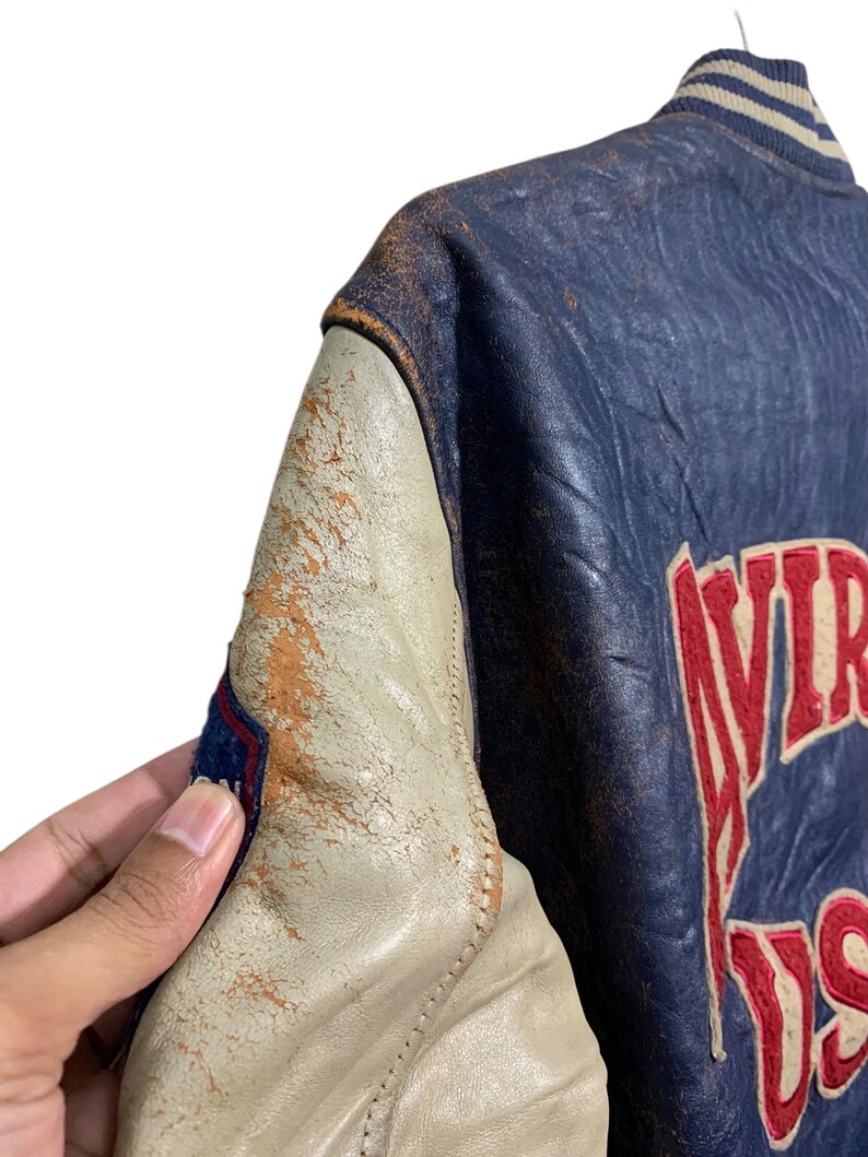 Puede incluir: Chaqueta universitaria vintage azul y crema con letras rojas. La chaqueta presenta un aspecto de cuero desgastado con cuello y pu&ntilde;os acanalados. El texto "VIRGINIA US" es visible en la parte posterior de la chaqueta.