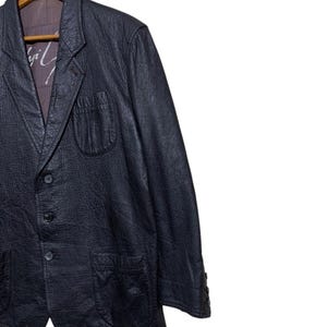 May include: A black leather blazer with a textured finish, featuring a notched lapel, three-button closure, and two front pockets. The blazer is hanging on a wooden hanger. The interior lining is visible, with a printed design.