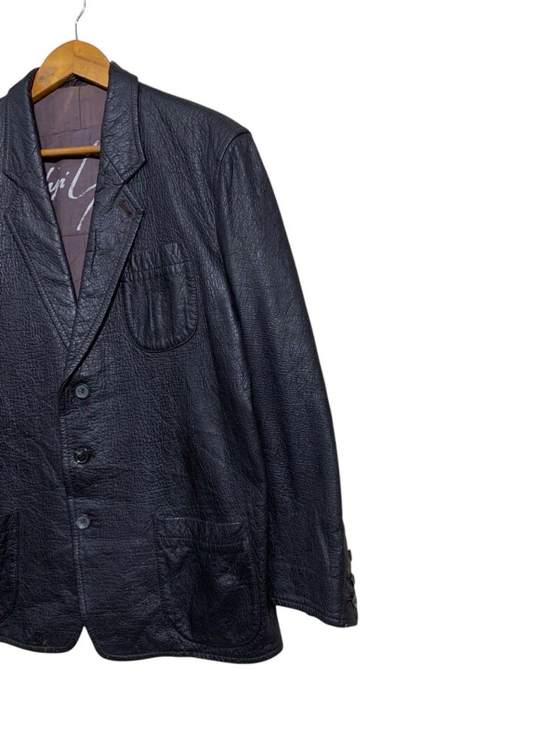 May include: A black leather blazer with a textured finish, featuring a notched lapel, three-button closure, and two front pockets. The blazer is hanging on a wooden hanger. The interior lining is visible, with a printed design.