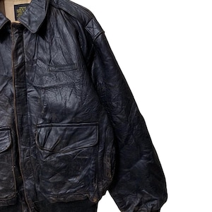 May include: A vintage black leather bomber jacket with a collared neckline, two front pockets, and a ribbed hem and cuffs. The jacket has a worn, distressed appearance, suggesting it is a pre-owned item. The jacket is hanging on a wooden hanger.