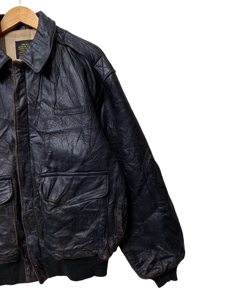 May include: A vintage black leather bomber jacket with a collared neckline, two front pockets, and a ribbed hem and cuffs. The jacket has a worn, distressed appearance, suggesting it is a pre-owned item. The jacket is hanging on a wooden hanger.