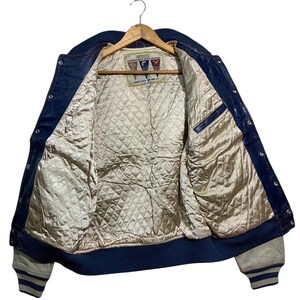 Puede incluir: Chaqueta de cuero azul vintage con forro beige acolchado, colgada de una percha de madera. La chaqueta presenta cuello, pu&ntilde;os y ribete azul marino, con pu&ntilde;os a rayas blancas y azules. La etiqueta interior es visible.