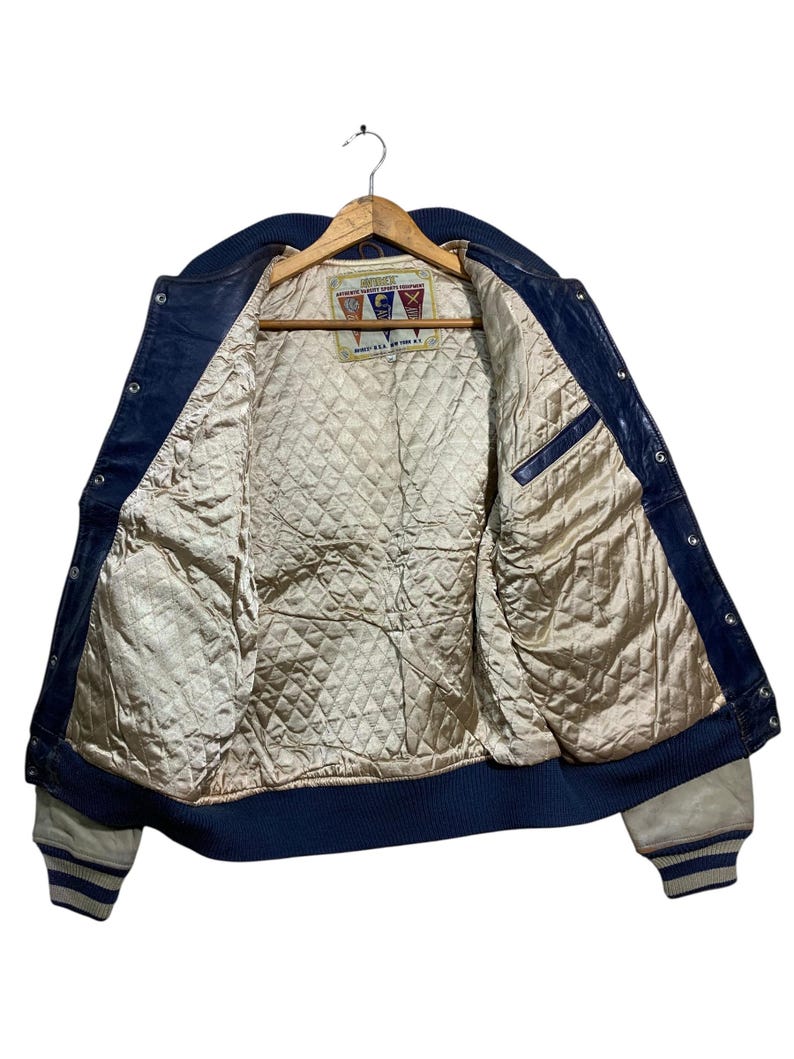 Puede incluir: Chaqueta de cuero azul vintage con forro beige acolchado, colgada de una percha de madera. La chaqueta presenta cuello, pu&ntilde;os y ribete azul marino, con pu&ntilde;os a rayas blancas y azules. La etiqueta interior es visible.