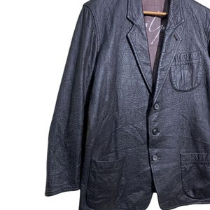May include: A black leather blazer with a textured finish. The jacket features a notched lapel, two front pockets, and a three-button closure. The interior lining is a contrasting color. The blazer is hanging on a wooden hanger.