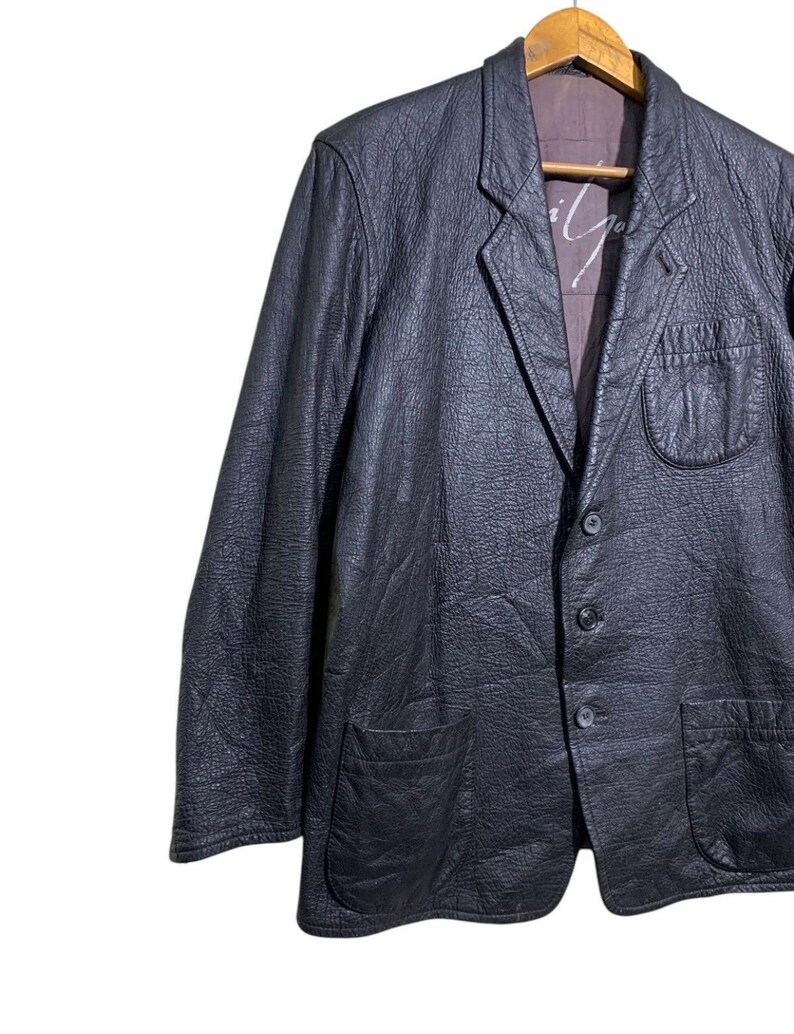 May include: A black leather blazer with a textured finish. The jacket features a notched lapel, two front pockets, and a three-button closure. The interior lining is a contrasting color. The blazer is hanging on a wooden hanger.