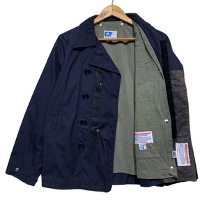 Puede incluir: Chaqueta azul marino de doble botonadura, con botones negros y un interior verde oliva en contraste. La chaqueta está colgada de una percha de madera, mostrando sus detalles y construcción.