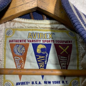 Puede incluir: Primer plano de la etiqueta interior de una chaqueta universitaria Avirex. La etiqueta muestra el logotipo de Avirex y el texto "Authentic Varsity Sports Equipment". Tambi&eacute;n muestra gr&aacute;ficos en forma de bander&iacute;n con las palabras "Chicago" y "Cleveland".
