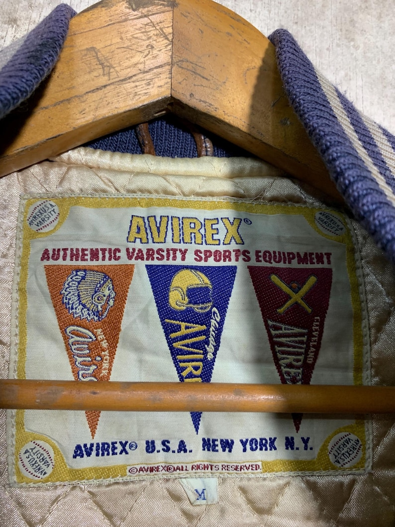 Puede incluir: Primer plano de la etiqueta interior de una chaqueta universitaria Avirex. La etiqueta muestra el logotipo de Avirex y el texto "Authentic Varsity Sports Equipment". Tambi&eacute;n muestra gr&aacute;ficos en forma de bander&iacute;n con las palabras "Chicago" y "Cleveland".
