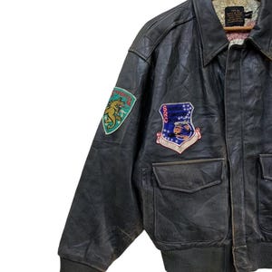 Może przedstawiać: Vintage czarna sk&oacute;rzana kurtka bomberka z naszywkami. Kurtka posiada dwie przednie kieszenie, prążkowany d&oacute;ł i mankiety oraz kołnierz. Naszywki obejmują zielonego i złotego smoka oraz niebiesko-czerwony emblemat z gwiazdami.