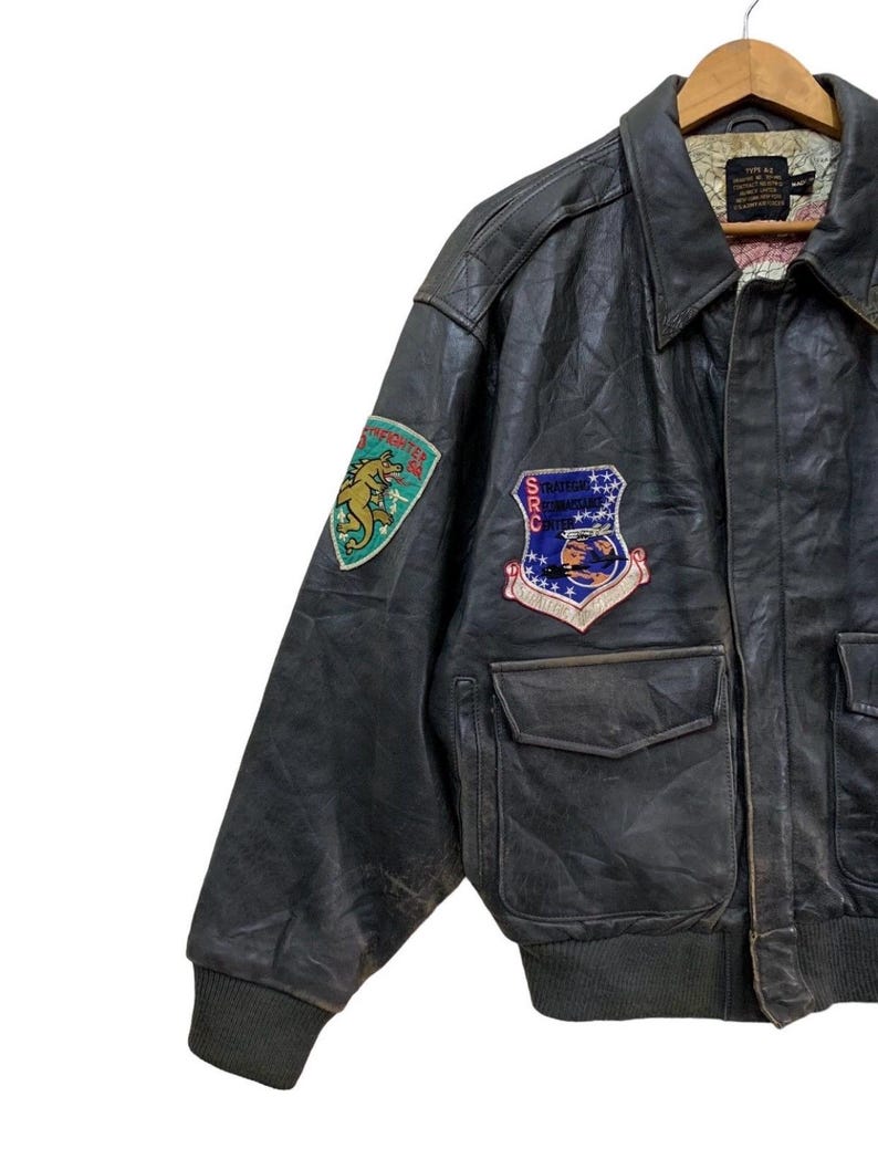 Może przedstawiać: Vintage czarna sk&oacute;rzana kurtka bomberka z naszywkami. Kurtka posiada dwie przednie kieszenie, prążkowany d&oacute;ł i mankiety oraz kołnierz. Naszywki obejmują zielonego i złotego smoka oraz niebiesko-czerwony emblemat z gwiazdami.
