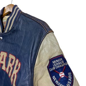 Puede incluir: Una chaqueta de b&eacute;isbol azul y crema vintage con la palabra "ARK" en letras grandes. La chaqueta tiene un parche de "Minor League Club Champion Newark Bears" en la manga. La chaqueta est&aacute; colgada en una percha de madera.