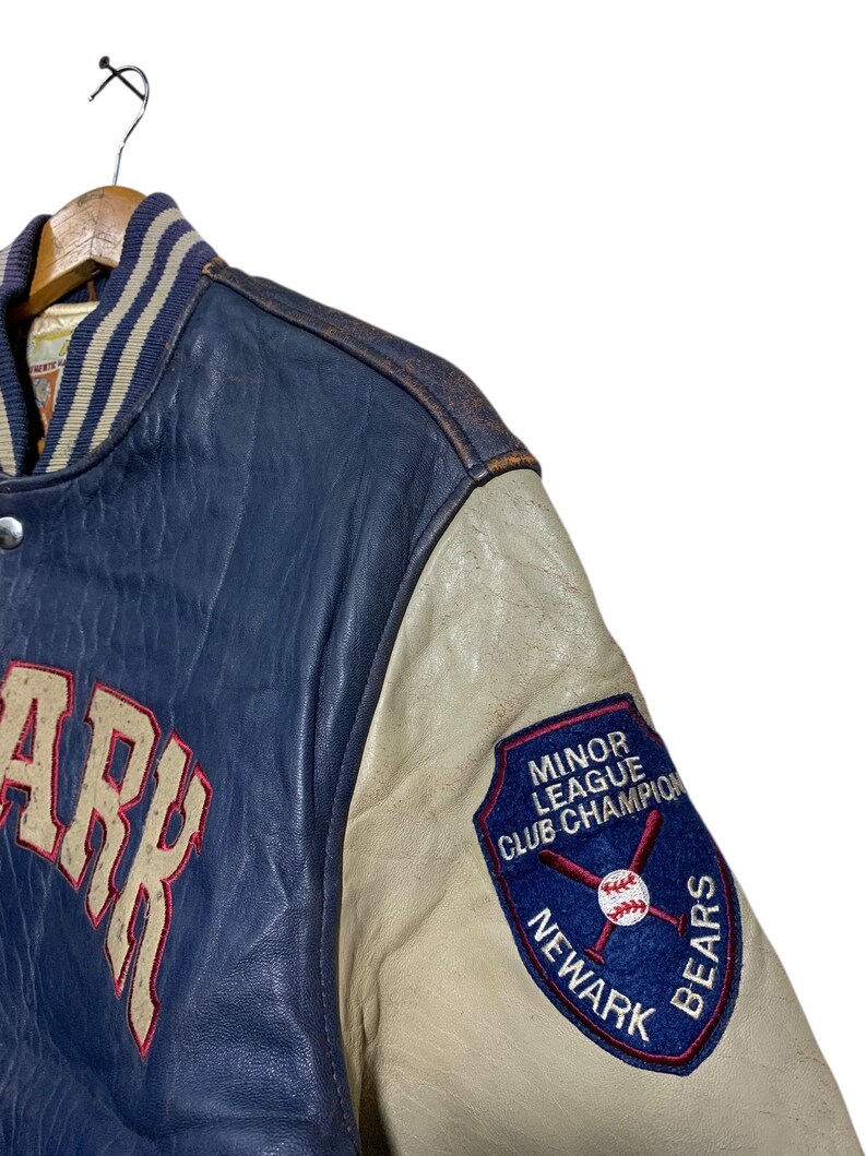 Puede incluir: Una chaqueta de b&eacute;isbol azul y crema vintage con la palabra "ARK" en letras grandes. La chaqueta tiene un parche de "Minor League Club Champion Newark Bears" en la manga. La chaqueta est&aacute; colgada en una percha de madera.