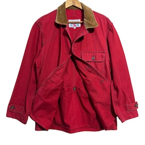 Puede incluir: Una chaqueta de algodón roja con cuello de pana marrón, cierre frontal de botones y un bolsillo en el pecho. La chaqueta incluye puños abotonados y una etiqueta en el interior del cuello con el texto "A" y "A" en un logo rojo y azul.