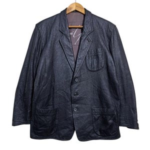 May include: A black leather blazer with a textured finish, featuring a notched lapel, three-button closure, and two front pockets. The blazer is hanging on a wooden hanger against a white background. The interior lining is visible.