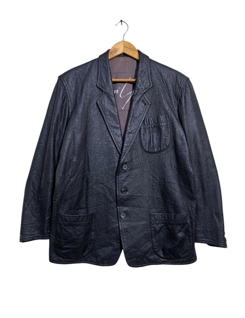 May include: A black leather blazer with a textured finish, featuring a notched lapel, three-button closure, and two front pockets. The blazer is hanging on a wooden hanger against a white background. The interior lining is visible.