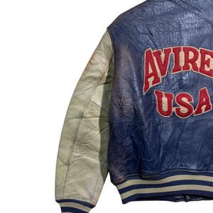 Puede incluir: Chaqueta de cuero vintage azul y crema con la palabra "AVIREX USA" en letras rojas en la espalda. La chaqueta tiene cuello y pu&ntilde;os acanalados con rayas azules y crema. La manga izquierda es de color crema.