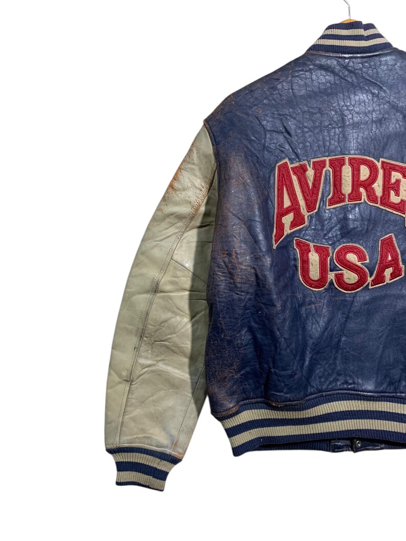 Puede incluir: Chaqueta de cuero vintage azul y crema con la palabra "AVIREX USA" en letras rojas en la espalda. La chaqueta tiene cuello y pu&ntilde;os acanalados con rayas azules y crema. La manga izquierda es de color crema.