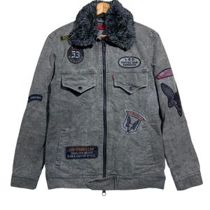 May include: Gray denim jacket with a faux fur collar and multiple patches. The jacket features a zip-up front, two chest pockets, and various embroidered patches with text and graphics. The Levi Strauss & Co. logo is visible.