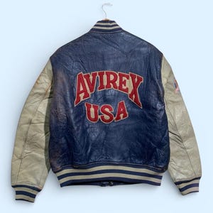 Puede incluir: Chaqueta de cuero azul y beige vintage con el texto "AVIREX USA" en letras rojas en la espalda. La chaqueta tiene mangas beige y pu&ntilde;os y cuello a rayas. La chaqueta est&aacute; colgada de una percha.