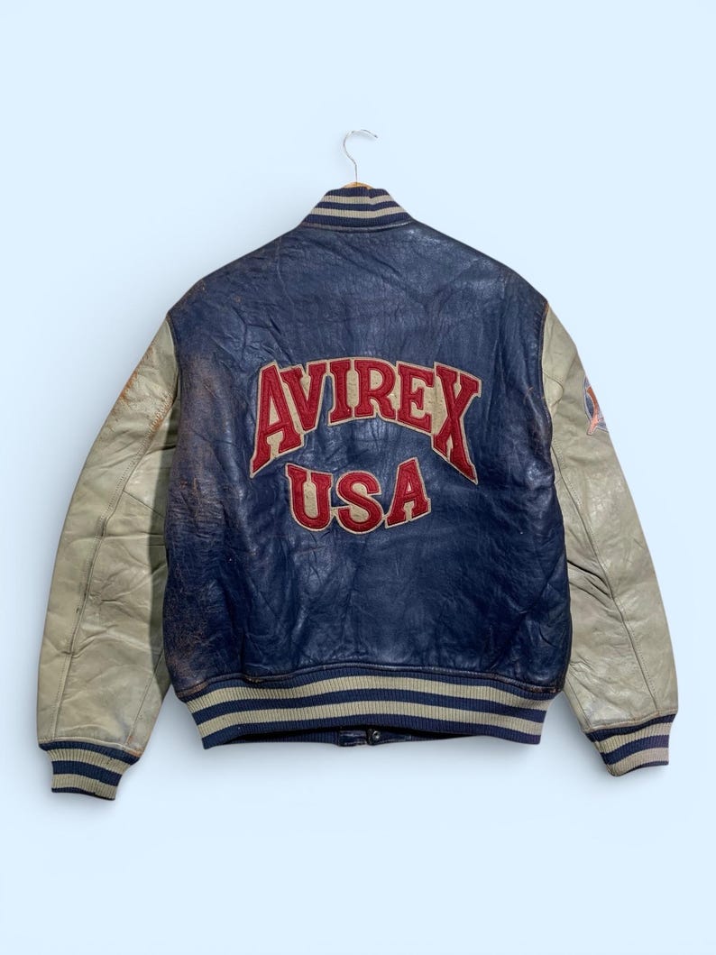 Puede incluir: Chaqueta de cuero azul y beige vintage con el texto "AVIREX USA" en letras rojas en la espalda. La chaqueta tiene mangas beige y pu&ntilde;os y cuello a rayas. La chaqueta est&aacute; colgada de una percha.