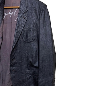 May include: Black leather blazer with a textured finish, featuring a notched lapel, a single-button closure, and a chest pocket. The interior lining is a muted purple with white text. The blazer is hanging on a wooden hanger.