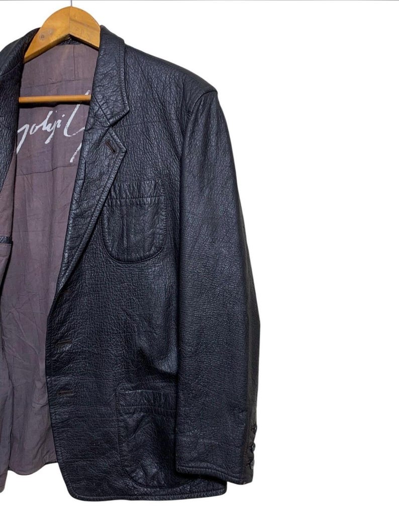 May include: Black leather blazer with a textured finish, featuring a notched lapel, a single-button closure, and a chest pocket. The interior lining is a muted purple with white text. The blazer is hanging on a wooden hanger.