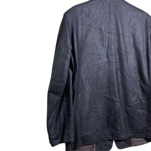 May include: A black leather jacket with a textured surface hangs on a wooden hanger. The jacket has a classic design with a notched collar and a button closure. The back of the jacket is visible.