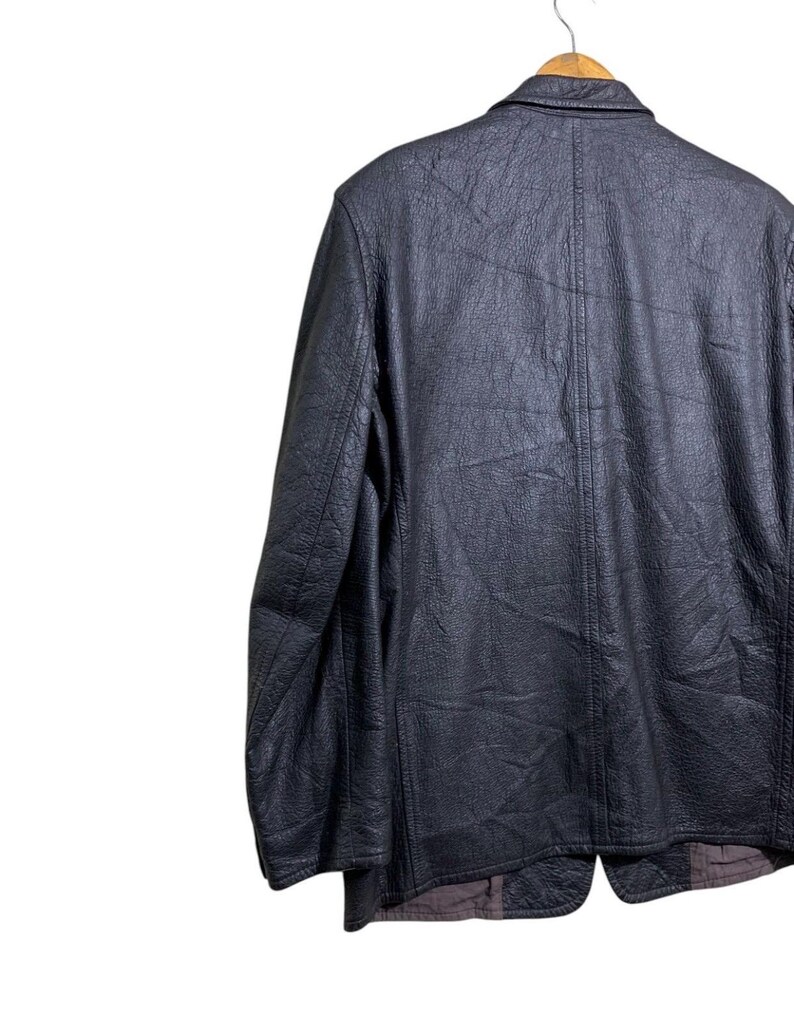May include: A black leather jacket with a textured surface hangs on a wooden hanger. The jacket has a classic design with a notched collar and a button closure. The back of the jacket is visible.