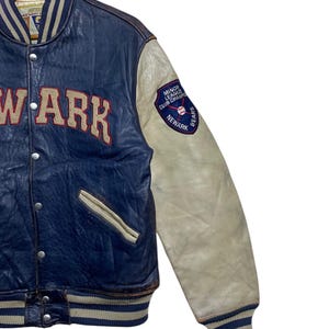 Puede incluir: Una chaqueta universitaria vintage de cuero azul y crema. La chaqueta presenta la palabra "NEWARK" en letras rojas grandes en el pecho. La manga derecha tiene un parche que dice "MINOR LEAGUE CLUB CHAMPS NEWARK BEARS".