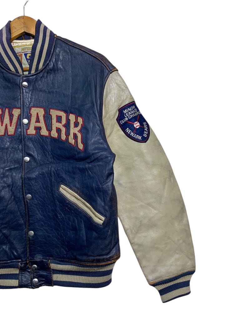 Puede incluir: Una chaqueta universitaria vintage de cuero azul y crema. La chaqueta presenta la palabra "NEWARK" en letras rojas grandes en el pecho. La manga derecha tiene un parche que dice "MINOR LEAGUE CLUB CHAMPS NEWARK BEARS".
