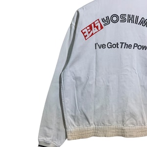 May include: White bomber jacket with black and white striped cuffs and collar. The back of the jacket features the text "YOSHIM" in black and red, with the phrase "I've Got The Power" below. The jacket has a ribbed hem.