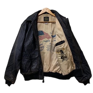 May include: Black leather bomber jacket with a ribbed collar and cuffs. The interior lining features an American flag, vintage aircraft illustrations, and a pin-up girl graphic. The jacket has two front pockets and a zipper closure.