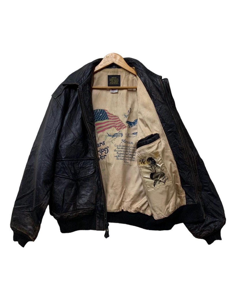 May include: Black leather bomber jacket with a ribbed collar and cuffs. The interior lining features an American flag, vintage aircraft illustrations, and a pin-up girl graphic. The jacket has two front pockets and a zipper closure.